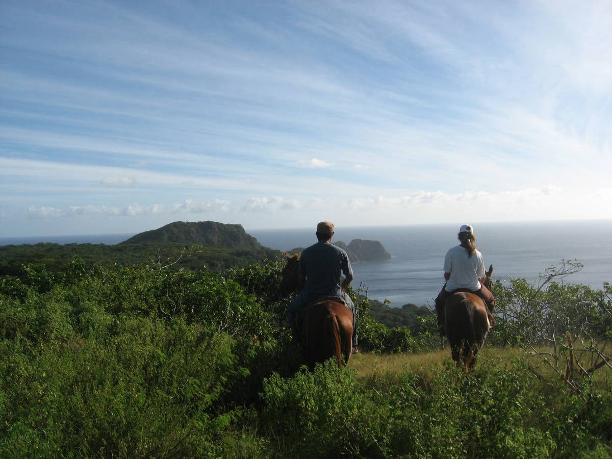 Um casal andando à cavalo em uma montanha em direção ao mar.